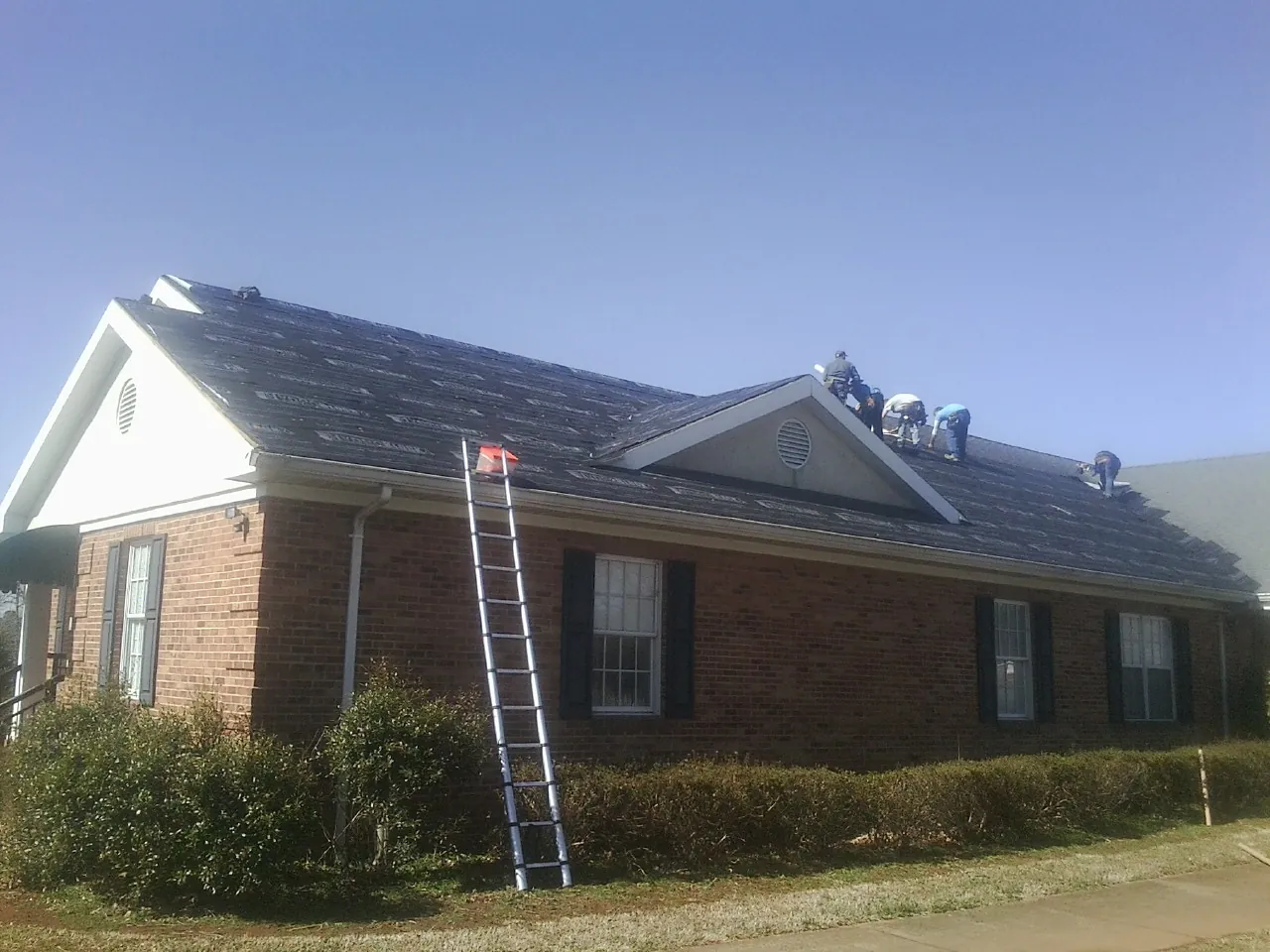 Crew tearing shingles off a brick ranch roof with a ladder set — Metal Roof Repair work by our crew in Gaston